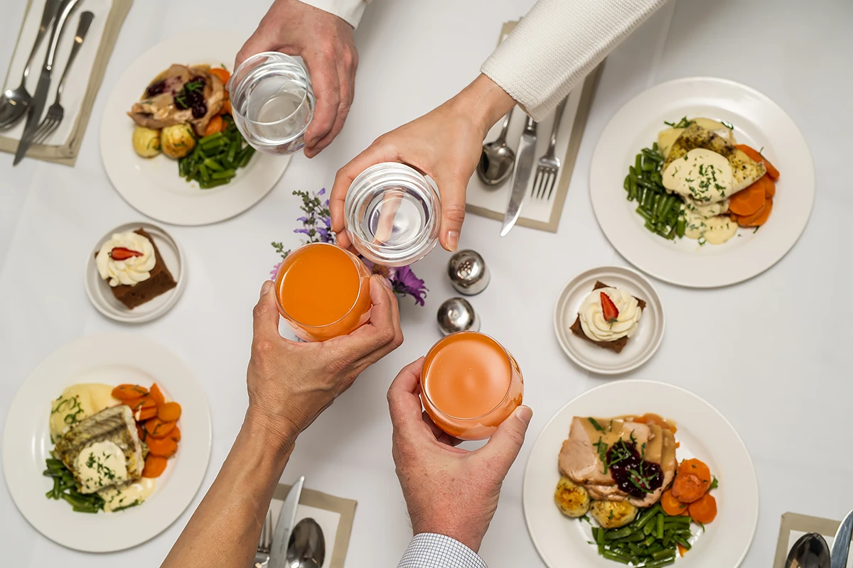 activities for seniors: An overhead shot of four hands making a toast with water and orange juice glasses over a beautifully set table with four plates of elegantly plated meals at a BASSCARE Boroondara Community Centre dining event.