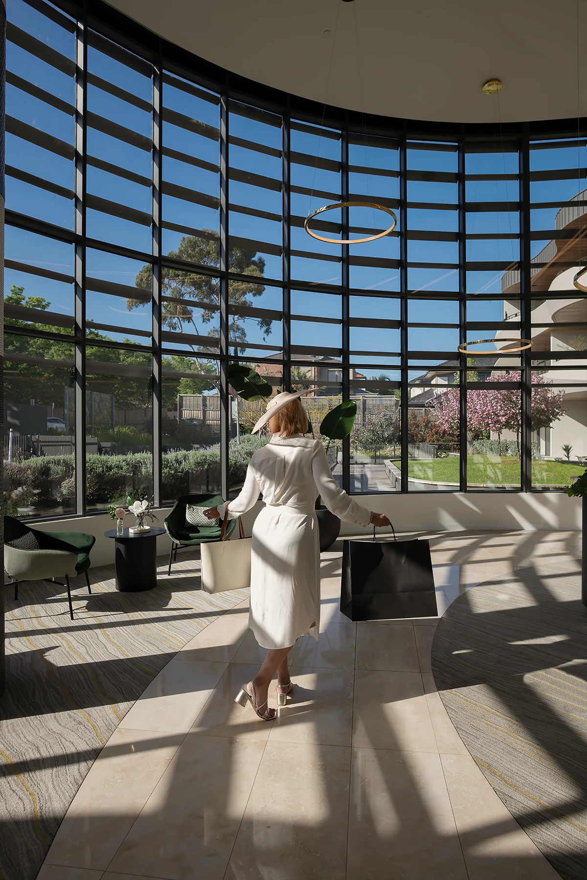 assisted living melbourne: A woman in an elegant white dress and wide-brimmed hat walks through the stunning, high-ceilinged atrium lobby of Morgan Glen Iris, carrying shopping bags after shopping at local Boroondara shops.