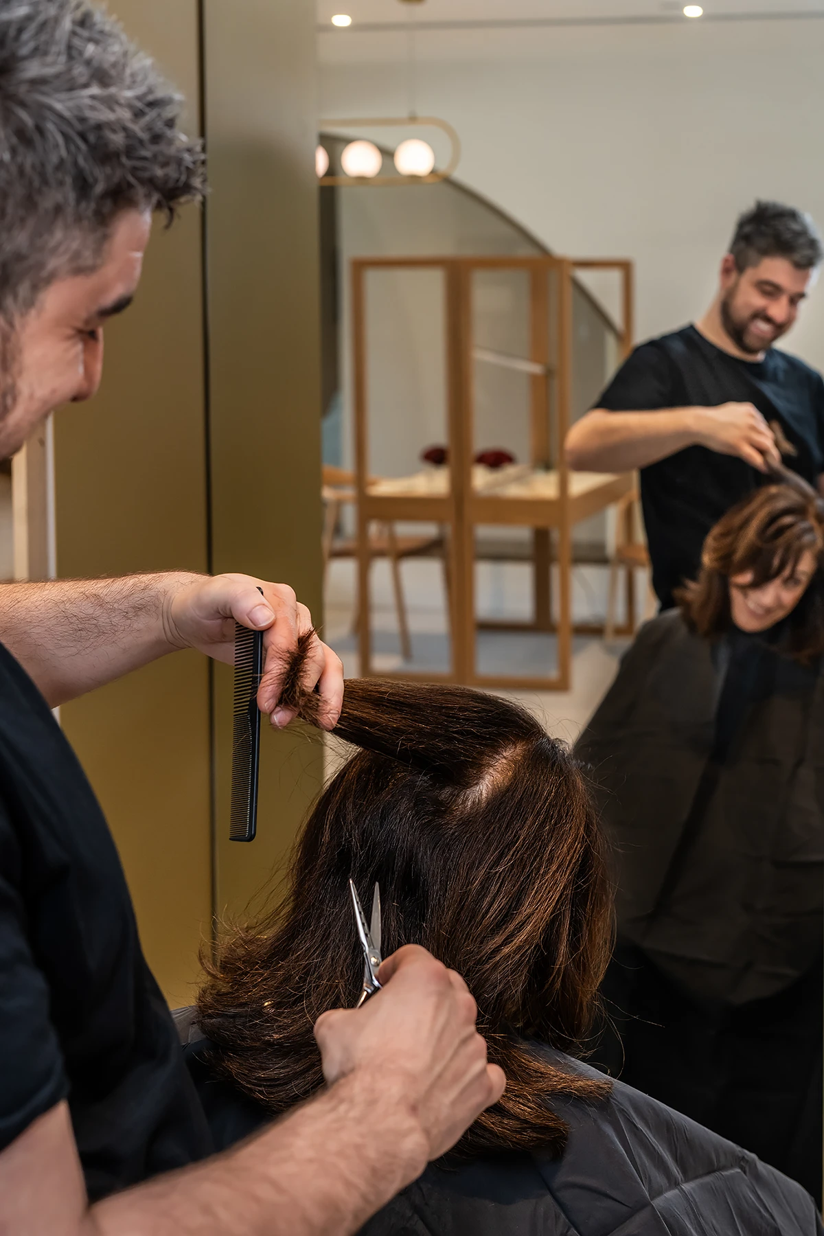assisted living melbourne: A skilled male hairstylist cutting a woman's brown hair in a bright, modern salon at Acacia Hair and Beauty at Morgan Glen Iris, with a reflection of the stylist smiling in the background.