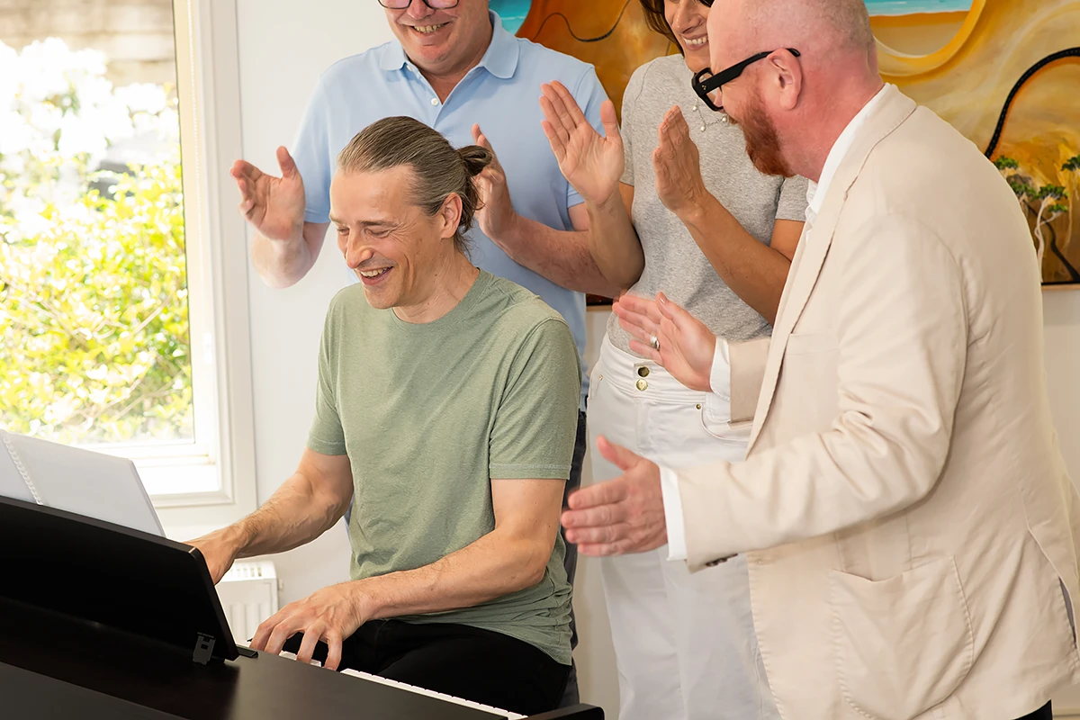 dementia support services: A group of four adults enjoying a BASSCARE Dementia Services music therapy session in Melbourne; a man in a green shirt is happily playing a black digital piano while three others clap and smile enthusiastically around him.