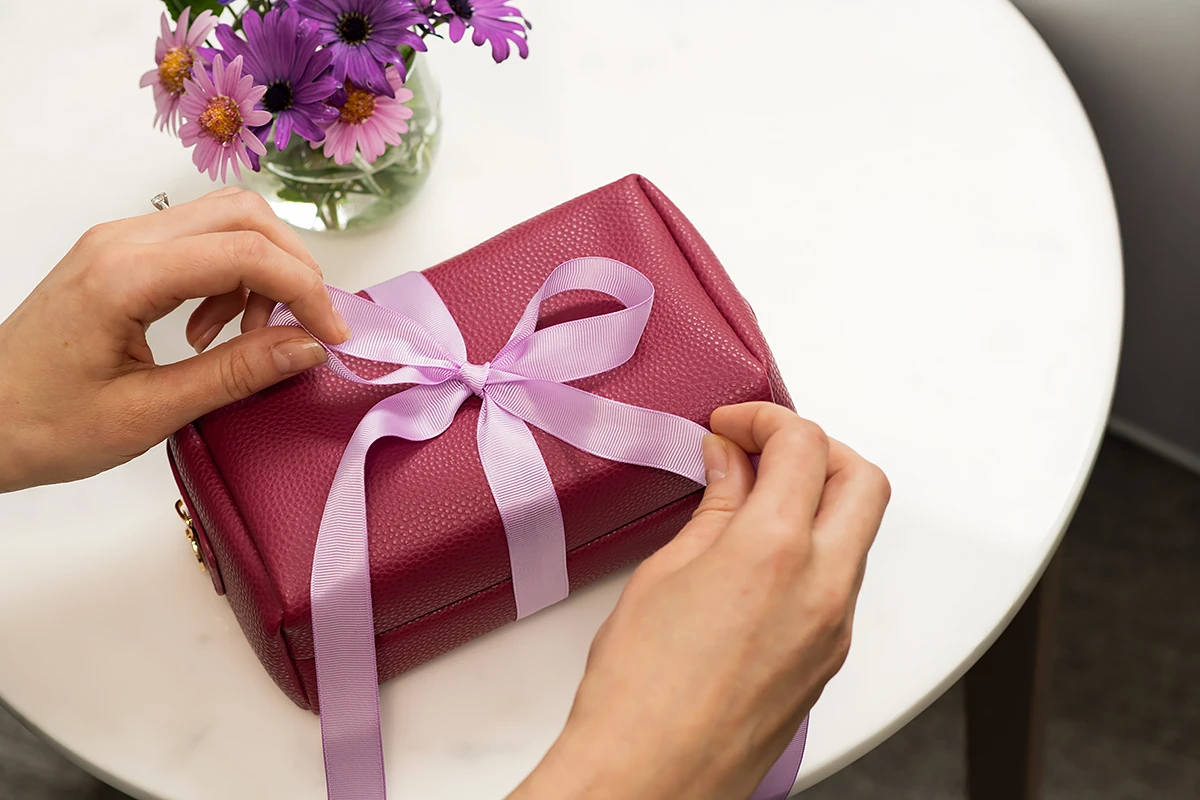 donate to BASSCARE: Close-up of a person's hands tying a light purple ribbon into a bow around a small, textured, magenta-coloured leather gift box, sitting on a table, encouraging a aged care donation.
