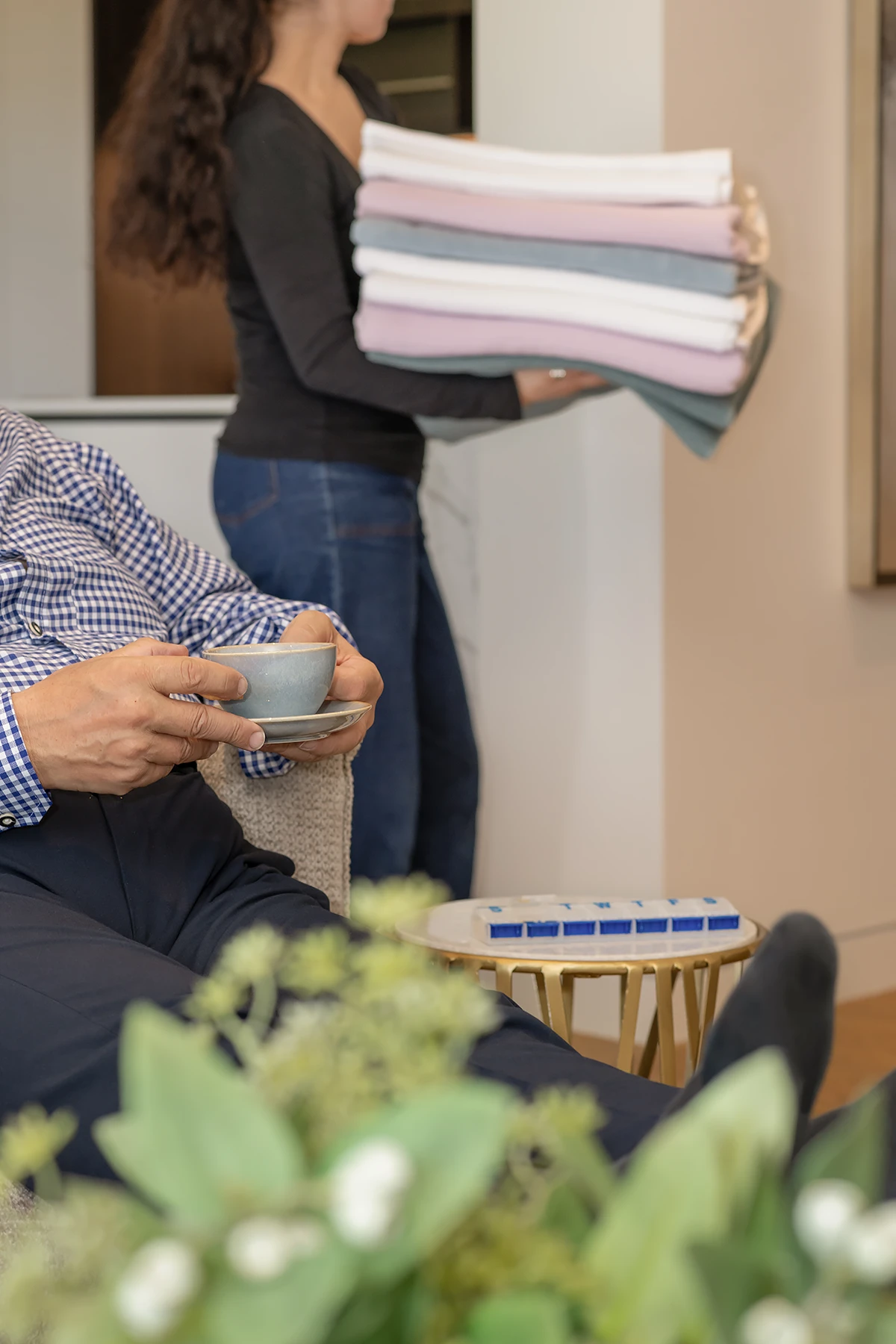 home care services: A client is relaxing with a cup of tea while a home care worker in the background carries a freshly folded stack of coloured laundry, illustrating home services support by BASSCARE in Boroondara.