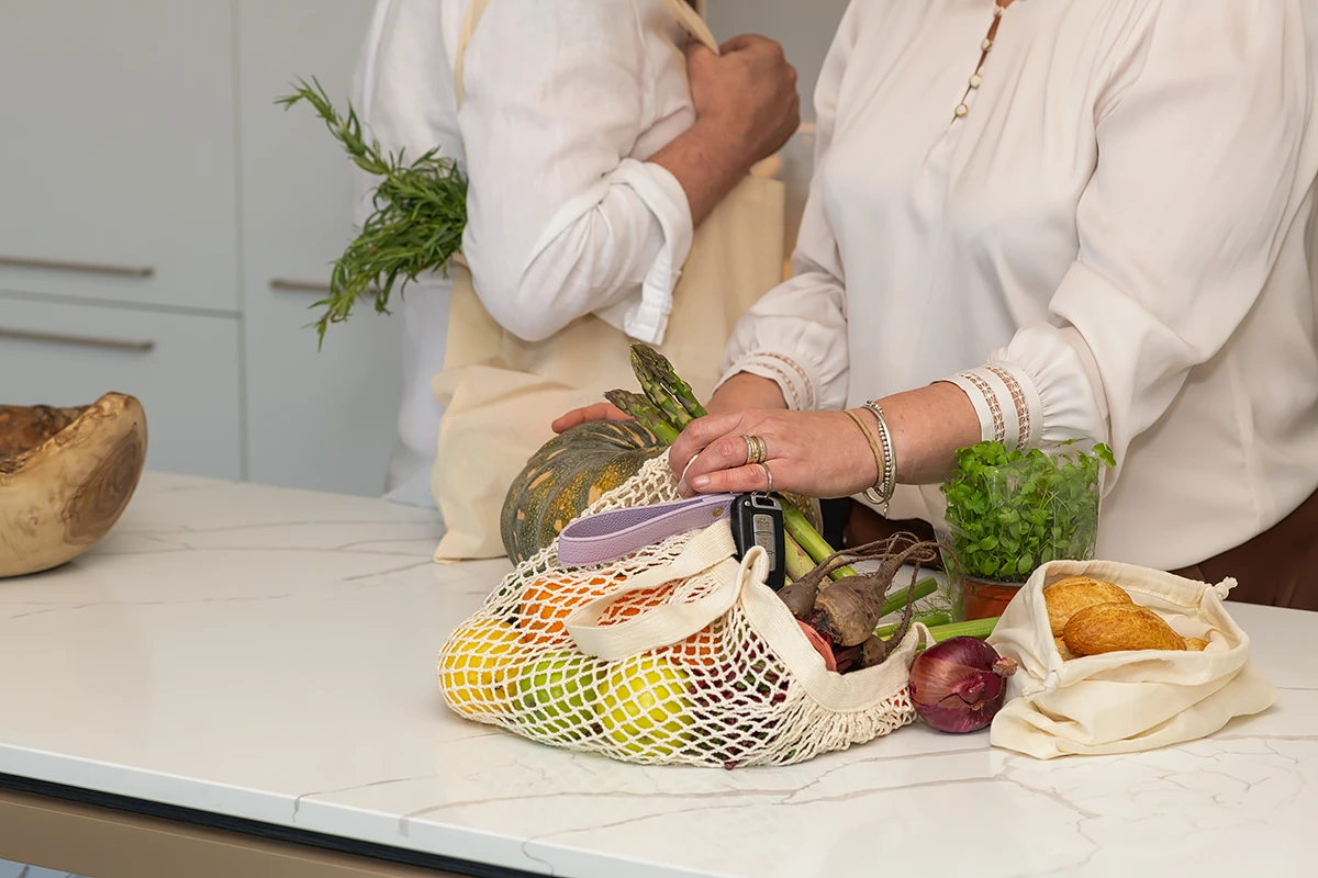 home care services: A person's hands placing a mesh bag full of fresh fruit and vegetables, including apples, carrots and beetroot, onto a kitchen bench after a melbourne home services shopping trip by BASSCARE.