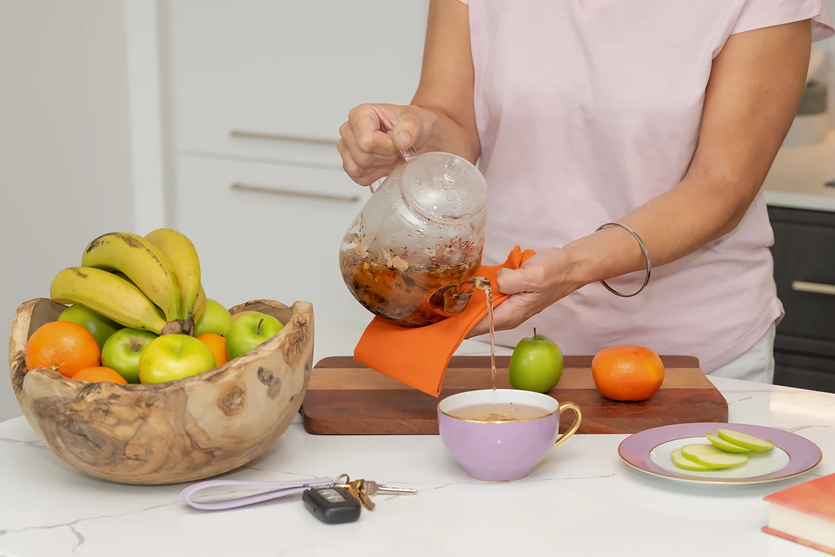 Independent Living Units: A close-up shot of a person pouring hot herbal tea from a glass teapot into a small purple teacup on a white kitchen counter, with a wooden bowl full of fruit nearby, conveying a Assisted Independent Living Unit homely lifestyle.