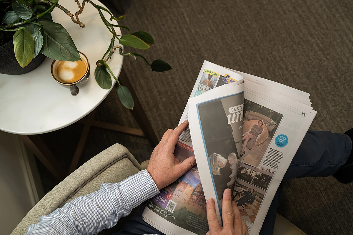 Residential Aged Care Ashburton: An overhead shot of a person relaxing in a Samarinda Lodge armchair, reading a broadsheet newspaper, with a cup of coffee on a nearby side table with a green houseplant.