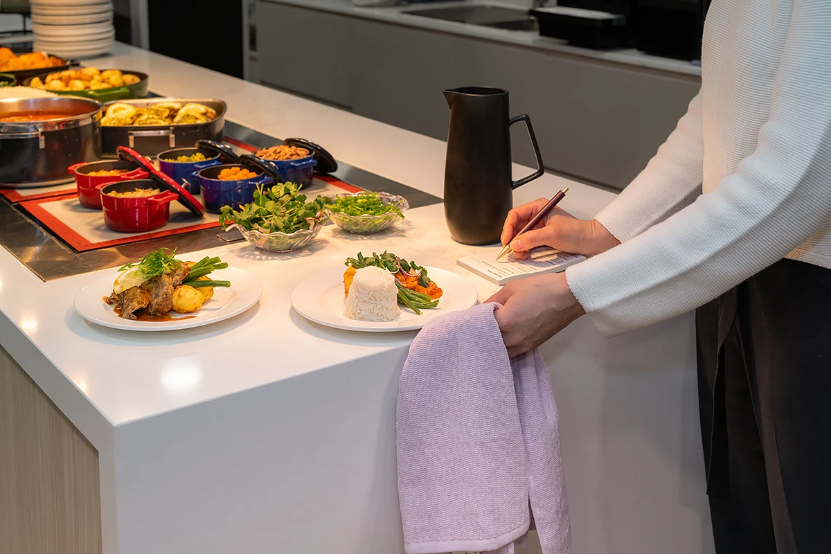 Residential Aged Care Canterbury: A BASSCARE hospitality staff member in a white textured sweater takes a note or order while standing at a bright service counter, which displays a variety of fresh and prepared meals in colorful bowls and on plates.