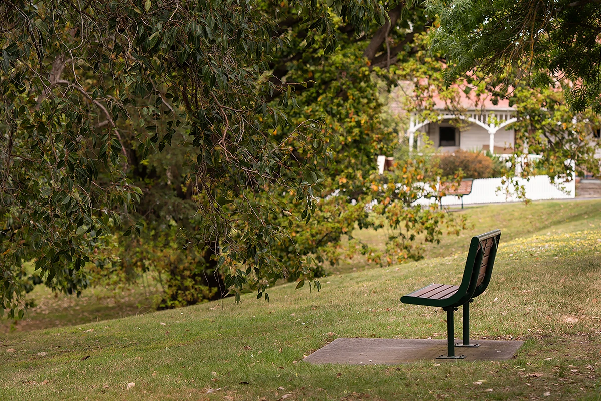 Retirement Living Melbourne: A tranquil view of a sloping grassy Boroondara park next to Hester Canterbury with a vacant wooden park bench in the foreground and a charming heritage house with a white verandah in the blurred background.